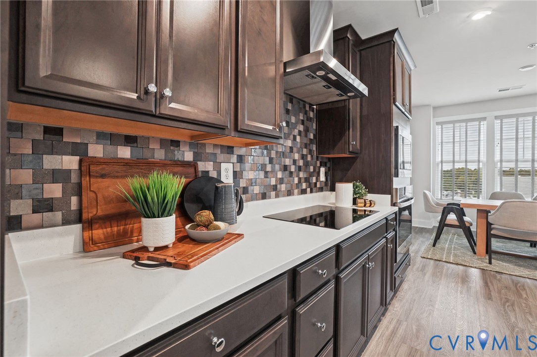 Kitchen featuring dark cabinetry, solid-surface countertops, mosaic tile backsplash, stainless steel range hood, and wood-finish flooring