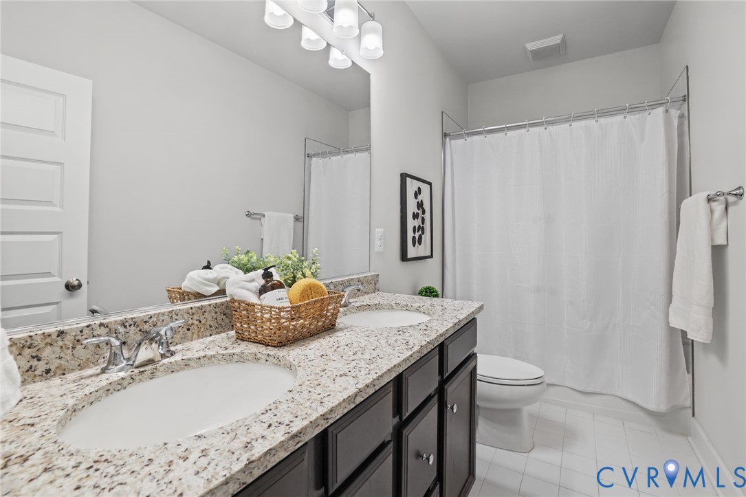 Bathroom featuring a double-sink vanity with stone-surfaced countertops, dark cabinetry, polished chrome fixtures, and a shower-tub combination with a white surround