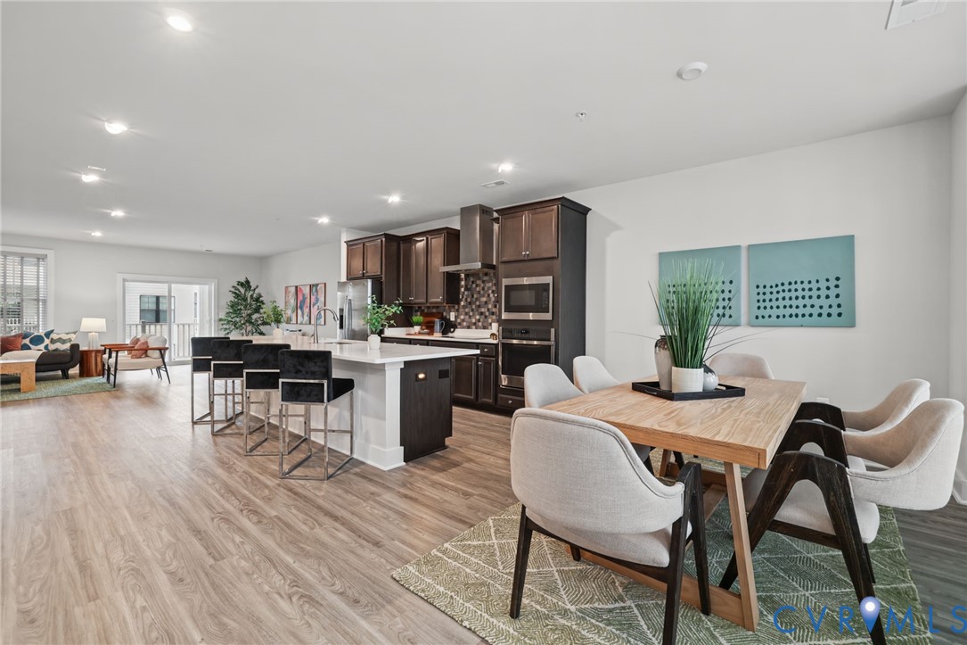 Kitchen featuring dark-finish cabinetry, quartz countertops with a waterfall edge, stainless steel built-in microwave and oven, stone tile backsplash, and wood-finish flooring