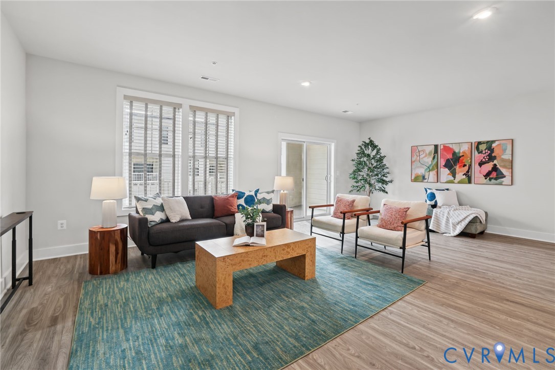Open-concept living space featuring wood-finish flooring, recessed lighting, double-paned windows with horizontal blinds, and a sliding glass door