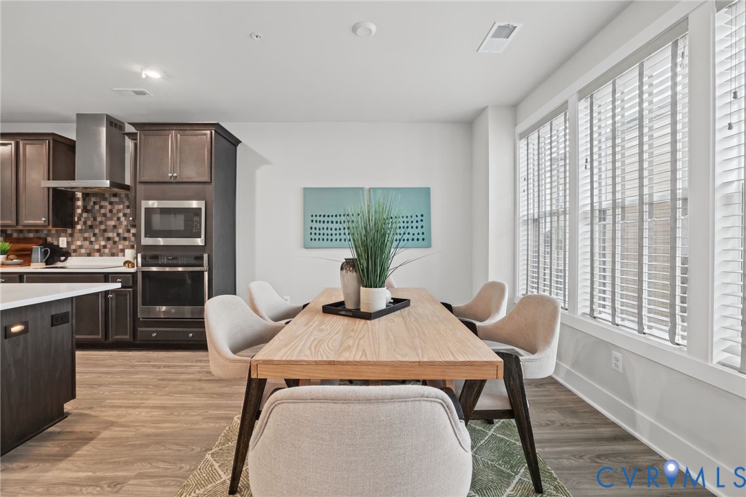 Open-concept area featuring wood-finish flooring, dark-toned cabinetry, stainless steel built-in oven, microwave, and expansive window coverage with horizontal blinds