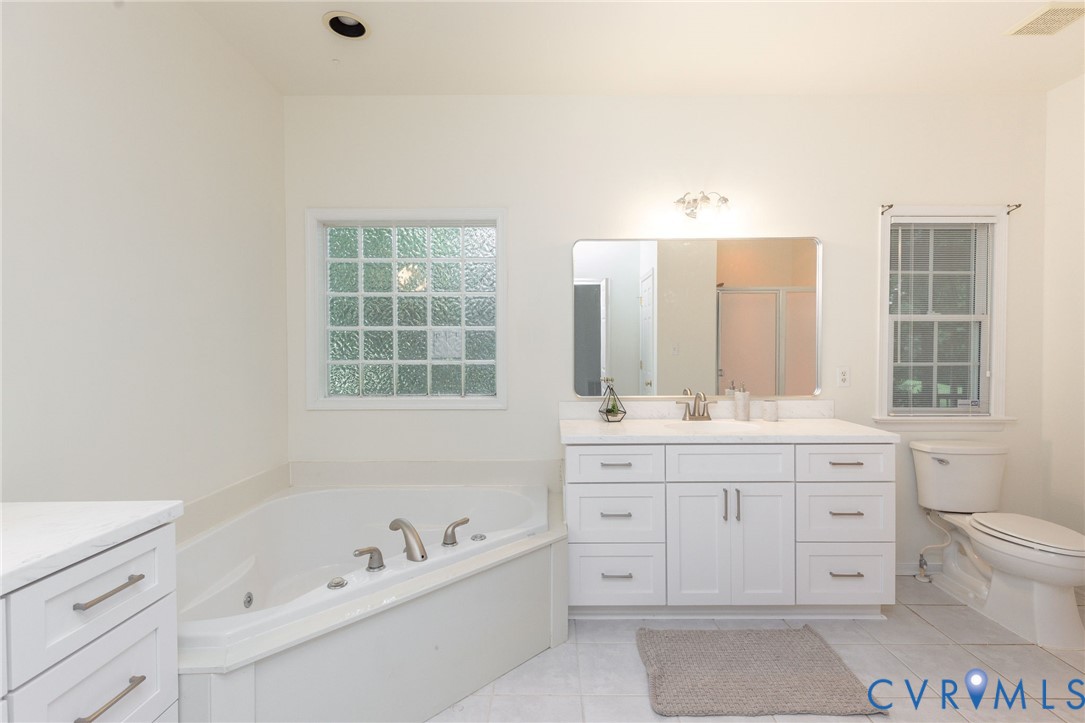 Corner soaking tub, white vanity with expansive countertop, large wall-mounted mirror, glass block window, and tile flooring