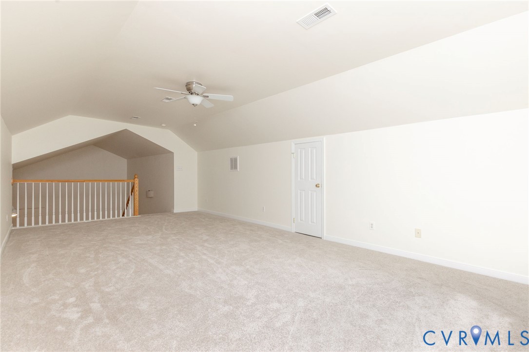 Vaulted ceiling featuring a central lighted ceiling fan, carpeted flooring, and a white wooden staircase railing