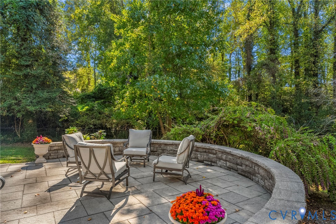 Curved stone retaining wall enclosing a paved patio area, set against a backdrop of mature trees