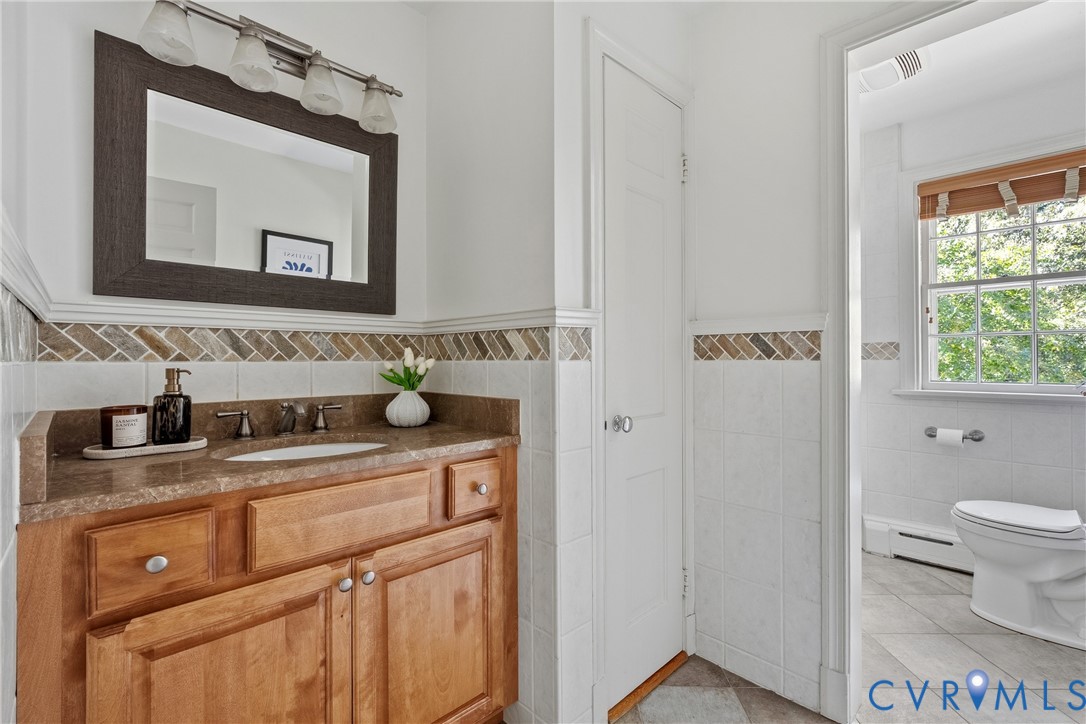 Bathroom featuring a wooden vanity, stone-finish countertop with an integrated sink, tri-light fixture, mirror with a dark frame, and decorative herringbone tile backsplash