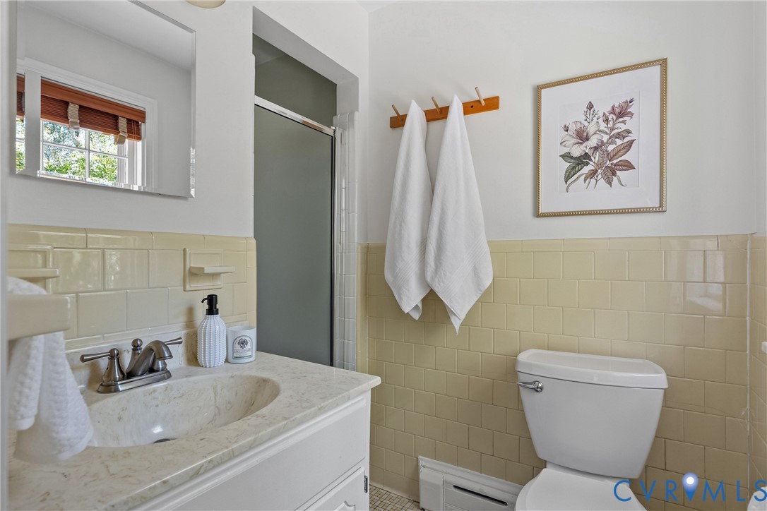 Bathroom featuring a vanity with stone-finish countertop, integrated oval basin, polished chrome faucet, partial-wall ceramic tile wainscoting, and a glass-enclosed shower