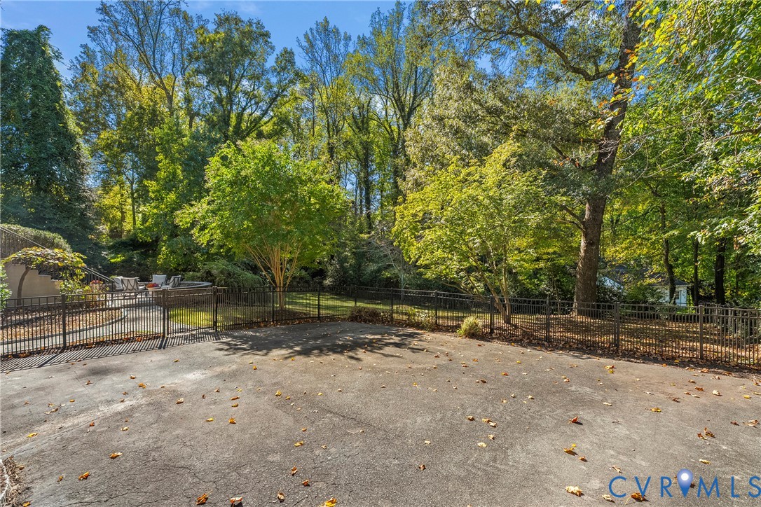 Spacious concrete patio enclosed by a metal perimeter fence, bordered by dense mature trees and varied deciduous foliage
