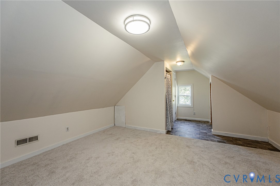 Attic space featuring vaulted ceilings, neutral wall tones, wall-to-wall carpeting, dual flush-mount light fixtures, and a multi-pane window