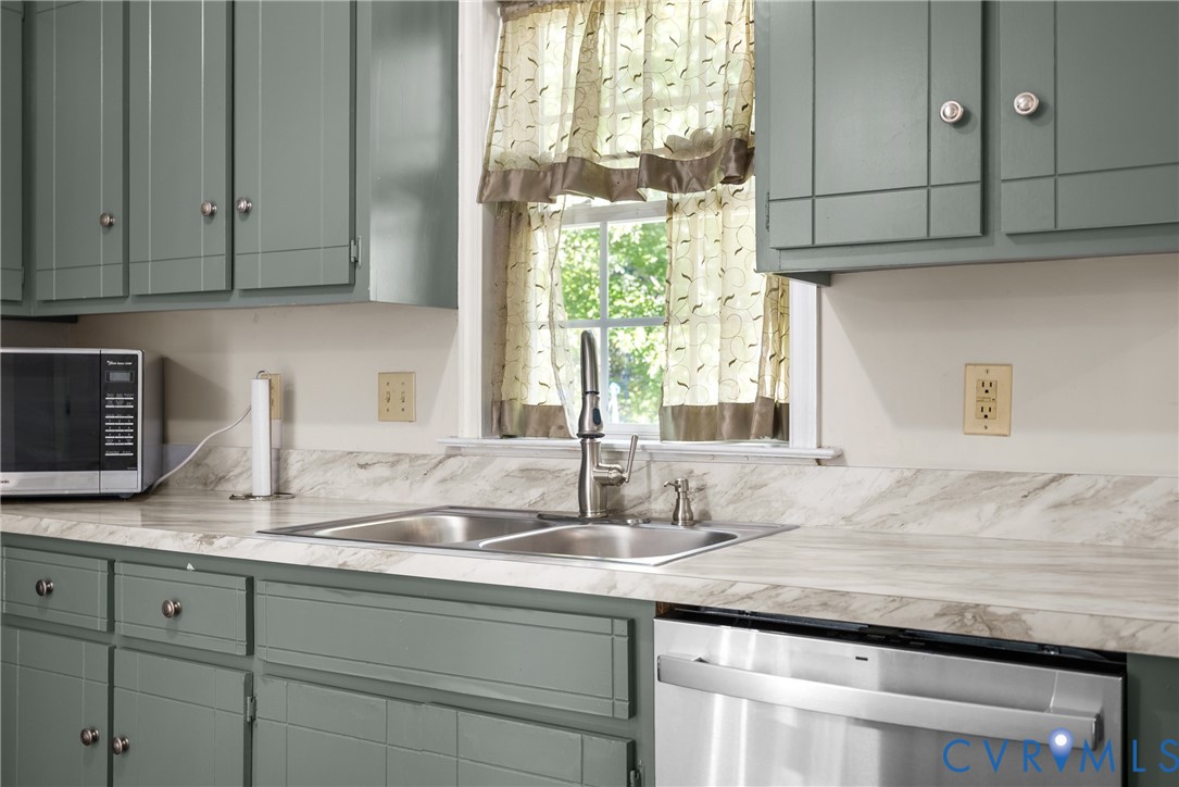 Kitchen featuring sage green paneled cabinetry, light-toned stone-patterned laminate countertops, dual-basin stainless steel sink, gooseneck faucet, and stainless steel dishwasher Kitchen featuring sage green paneled cabinetry, light-toned stone-patterned laminate countertops, dual-basin stainless steel sink, gooseneck faucet, and stainless steel dishwasher