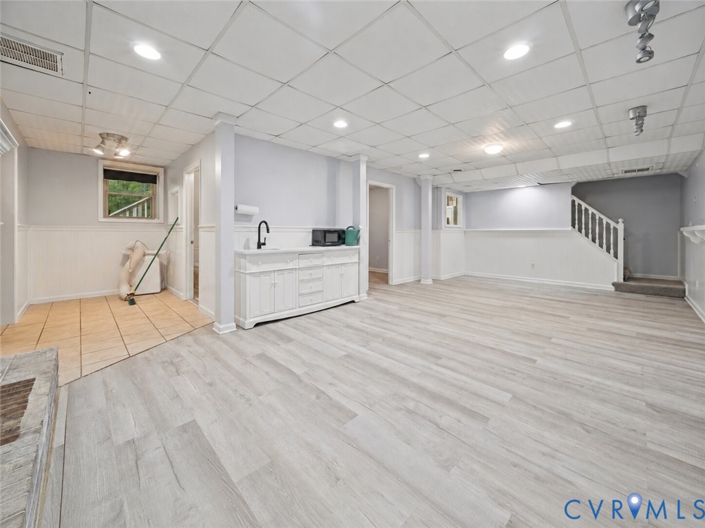 Open-concept area featuring wood-finish flooring, white built-in cabinetry with a sink, drop ceiling with recessed lighting, beadboard wainscoting, and a carpeted staircase with white balusters
Virtually edited