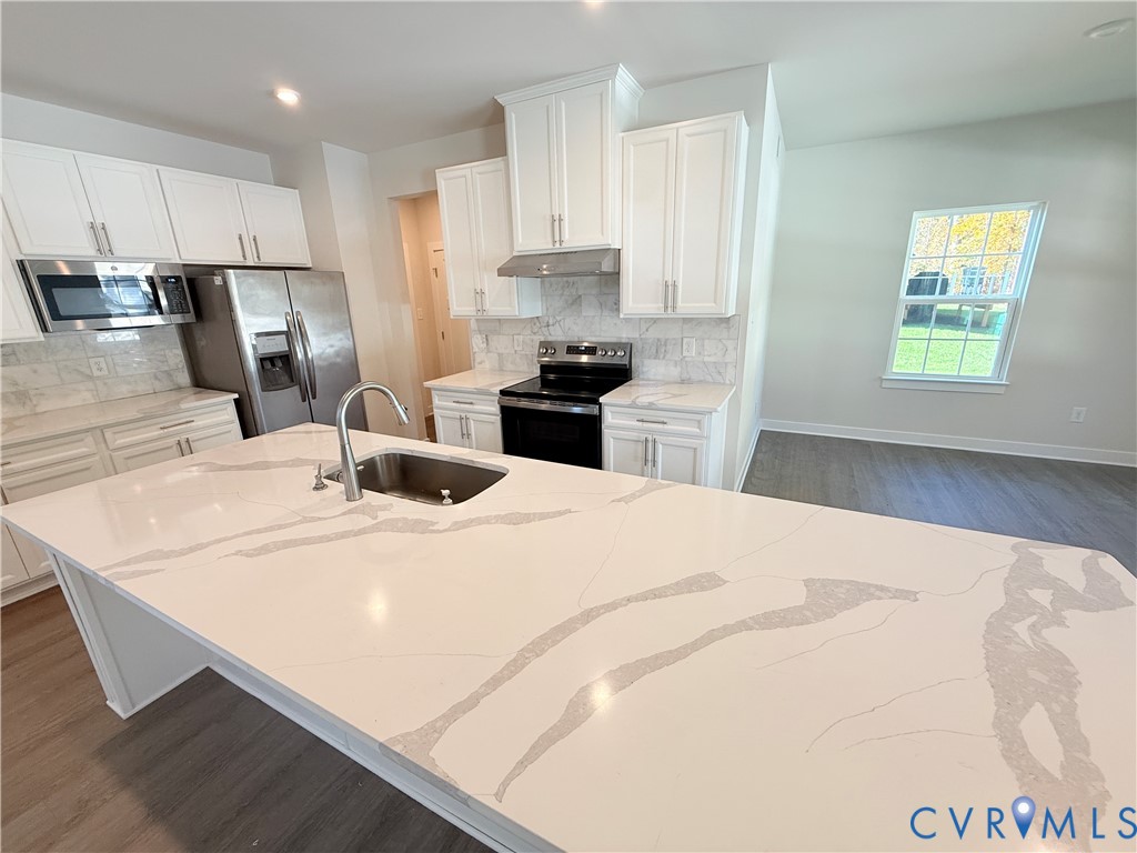 Kitchen featuring white shaker-style cabinetry, a large central island with a solid surface countertop, stainless steel appliances, stone-look tile backsplash, and wood-finish flooring