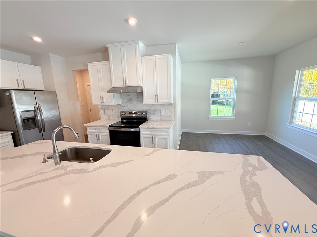 Kitchen featuring white shaker-style cabinetry, a quartz countertop island with an undermount sink, stone-finish backsplash, stainless steel appliances, and wood-finish flooring