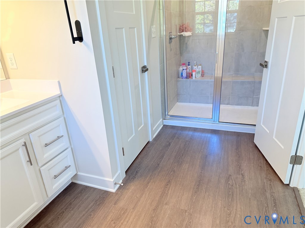 White vanity with brushed metal hardware, walk-in shower with a built-in bench, neutral tile shower surround, glass shower enclosure, and wood-finish flooring