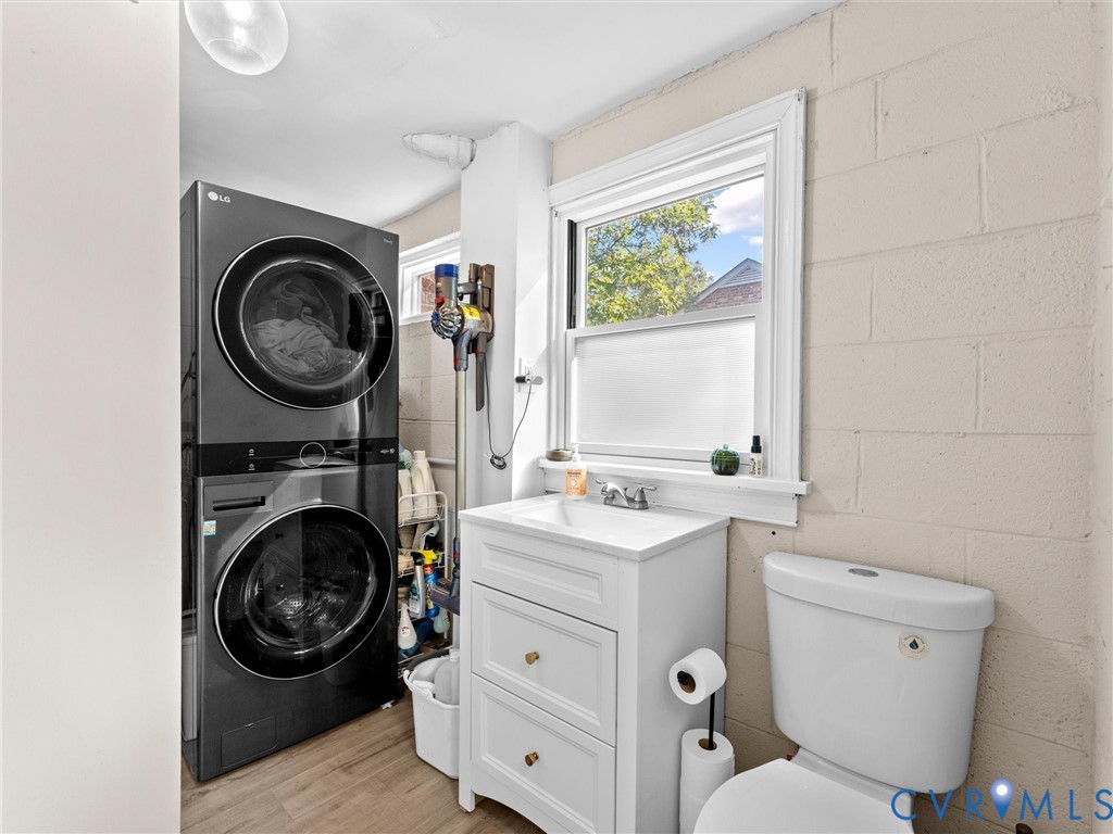 White vanity with a rectangular sink, dual-stack front-load washer and dryer, wood-finish flooring, and painted masonry walls