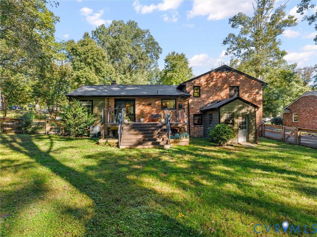 Multi-level brick exterior featuring a raised wood deck with staircase, expansive lawn, and fenced perimeter