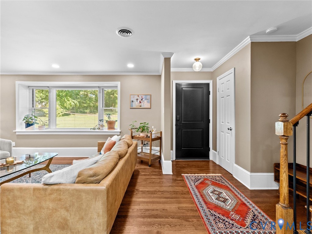 Living area featuring wood-finish flooring, a large bay window, crown molding, a solid-panel door, and a wooden handrail with balusters