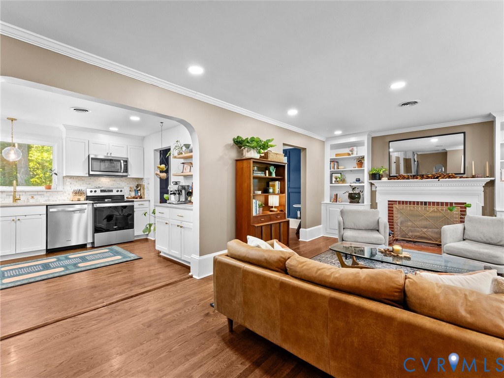 Open-concept kitchen featuring white cabinetry, stainless steel appliances, and wood-finish flooring, centered by an arched transition to the living area with a fireplace and built-in shelving