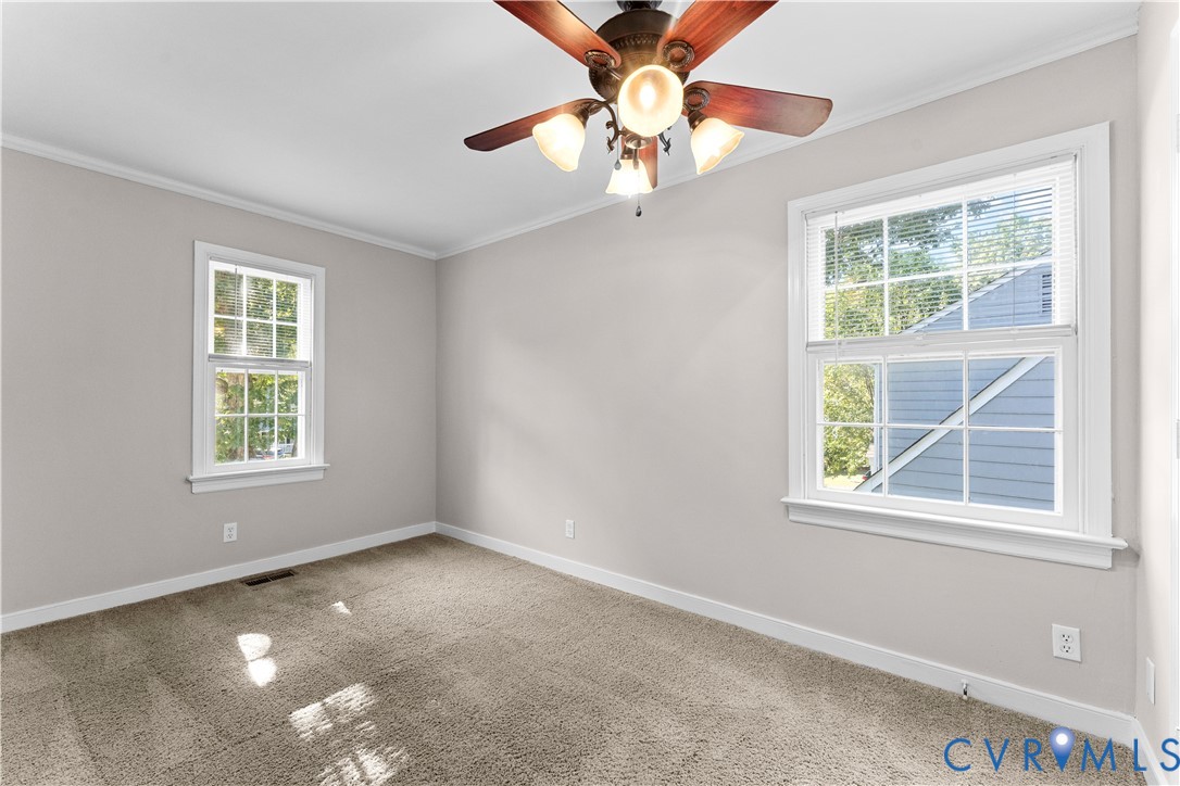 Neutral interior featuring neutral-toned carpeting, two windows with white casings, and a ceiling fan with four integrated light fixtures