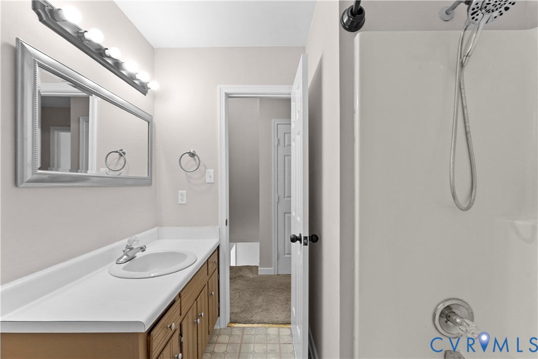 Bathroom featuring a vanity with a white integrated countertop and sink, large wall-mounted mirror, multi-bulb overhead light fixture, and a shower with a handheld showerhead