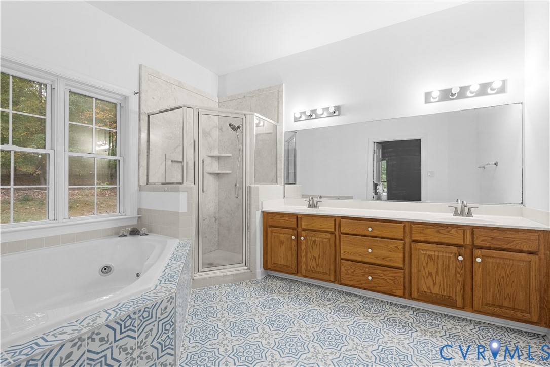 Dual-sink vanity with wood-finish cabinetry, corner soaking tub, glass-enclosed shower, patterned tile flooring, and a full-width wall mirror