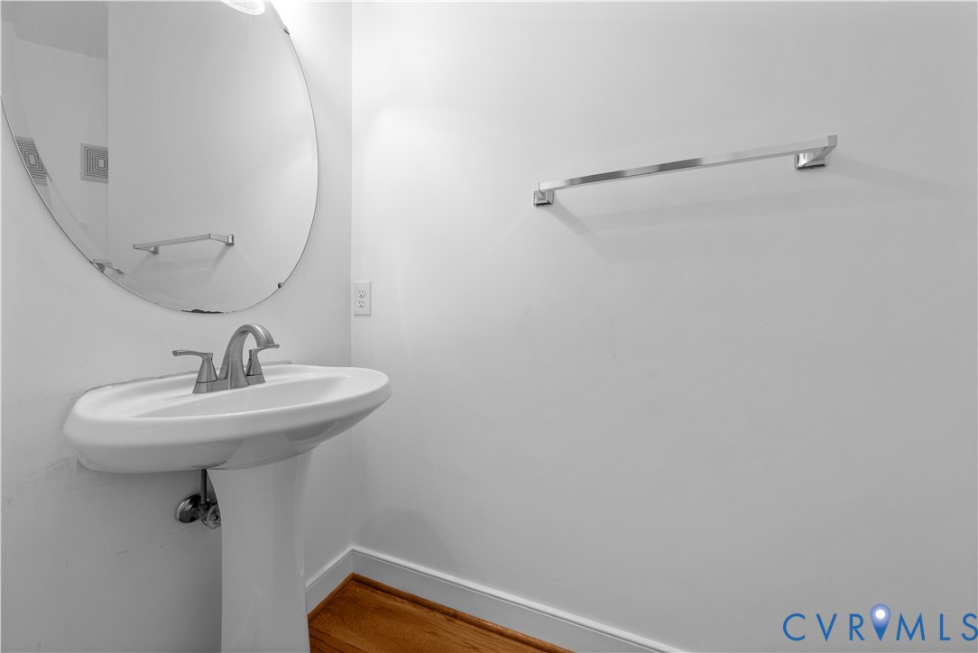 Pedestal sink featuring a polished chrome faucet, oval wall-mounted mirror, and wall-mounted towel bar over wood-finish flooring