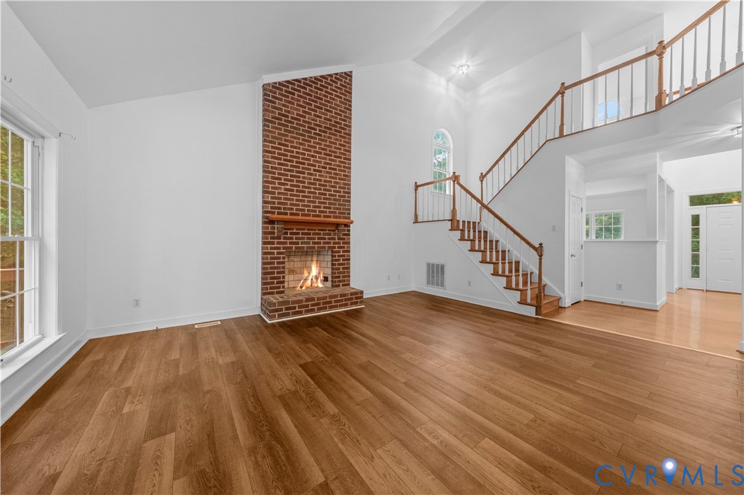 Vaulted ceiling, floor-to-ceiling brick fireplace with wood mantel, wood-finish flooring, and staircase featuring a wooden handrail and balusters