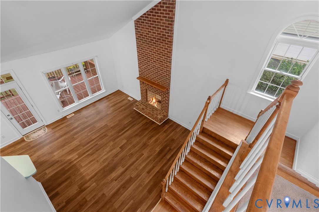 Vaulted ceilings, floor-to-ceiling brick fireplace with a wood mantel, wood-finish flooring, staircase with wood handrails and white balusters, and arched windows