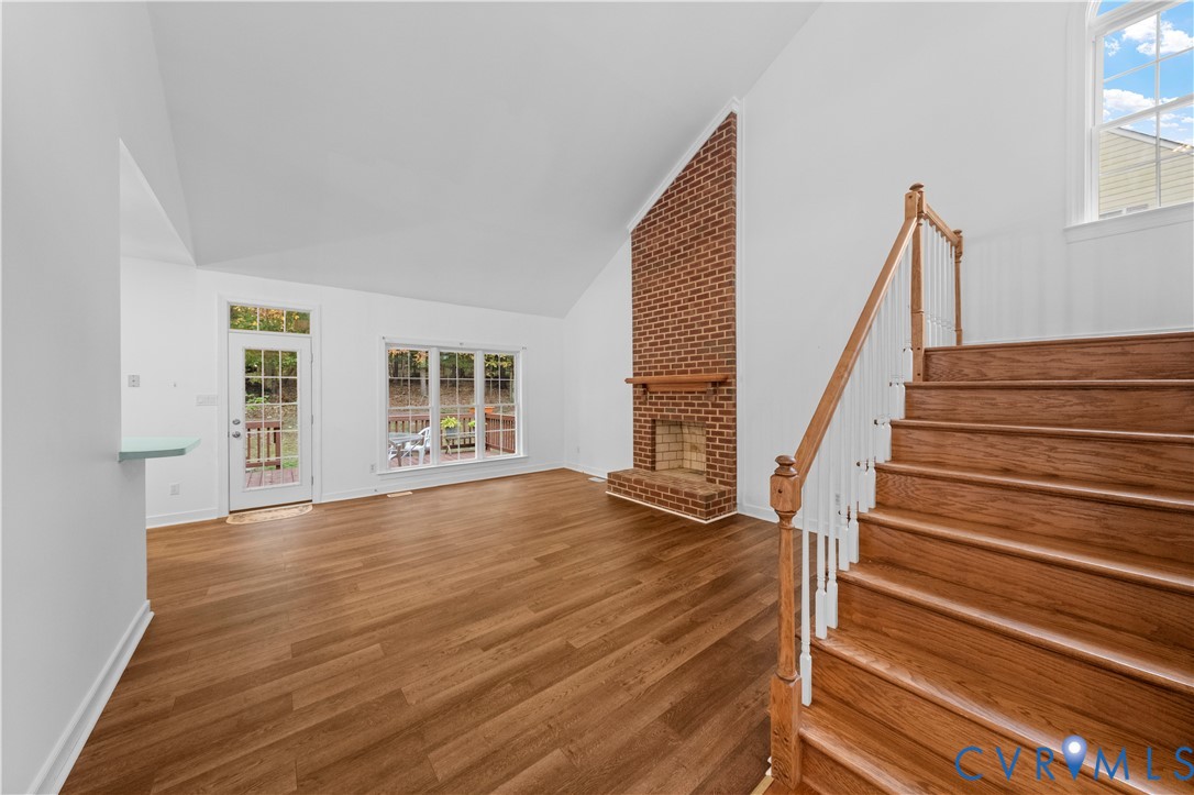 Living area featuring a vaulted ceiling, floor-to-ceiling brick fireplace with wood mantel, wood-finish flooring, and a staircase with a wood handrail