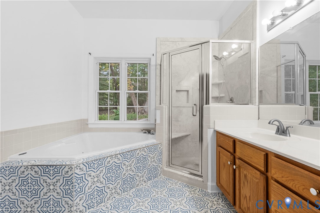 Bathroom featuring a jetted tub with patterned tile surround, walk-in glass shower with stone-finish walls, double-door wood cabinetry, and large-format wall mirror