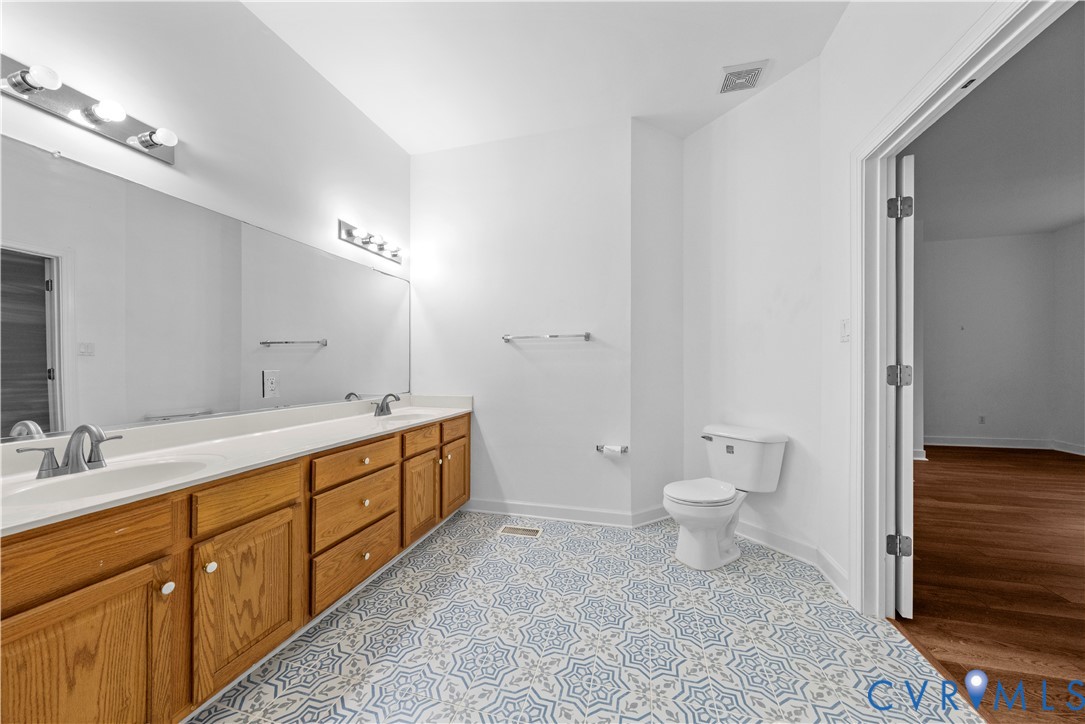 Dual-sink vanity with wood-finish cabinetry, countertop-integrated sinks, full-width vanity mirror, multi-bulb wall lighting, and patterned tile flooring