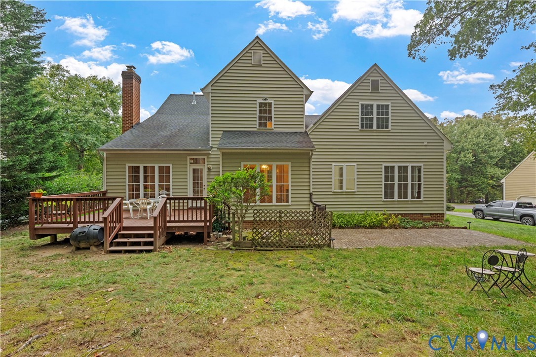 Two-story residence with light-colored horizontal siding, a multi-gabled roofline, a raised wooden deck, a paved stone patio, and an external brick chimney