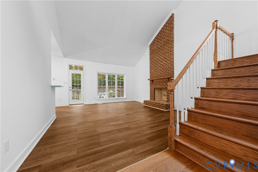 Vaulted ceiling, floor-to-ceiling brick fireplace with wood mantel, wood-finish flooring, and staircase with wooden handrails and balusters