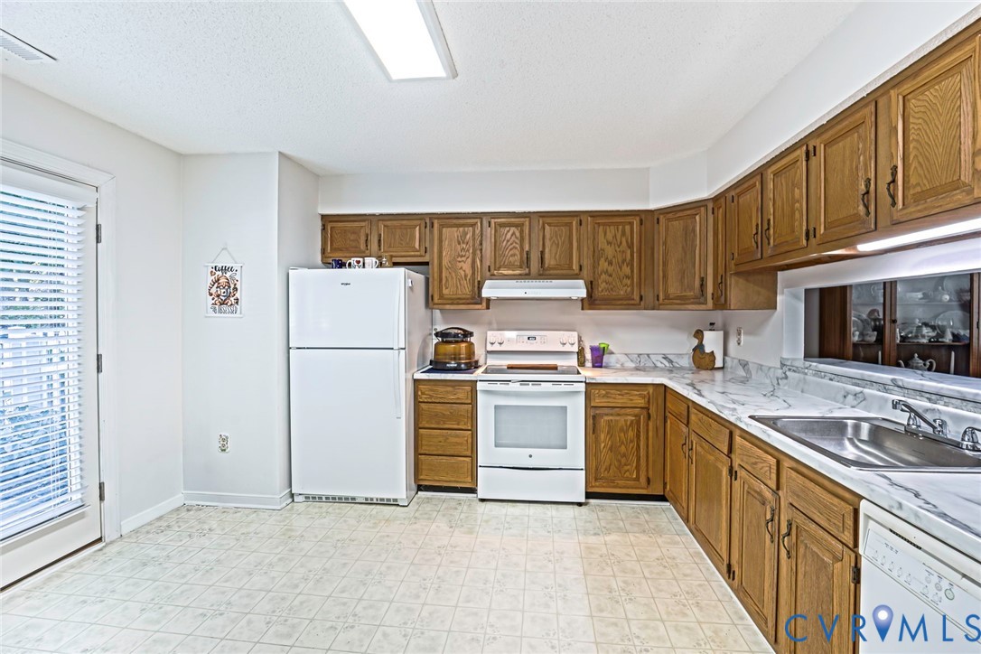 Kitchen featuring natural wood cabinetry, stone-patterned countertops, stainless steel sink, and white appliances including a built-in dishwasher and range.