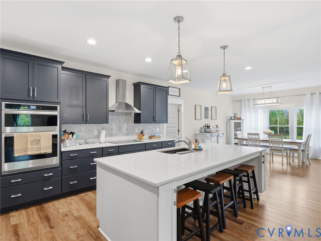 Kitchen featuring charcoal cabinetry, central island with solid-surface countertop, stainless steel double wall oven, stainless steel range hood, and patterned tile backsplash