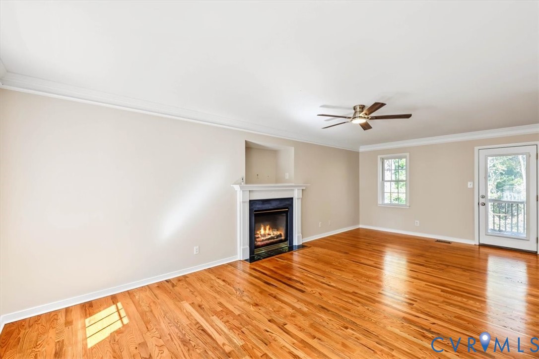 Living area featuring wood-finish flooring, a fireplace with a white mantel, crown molding, a ceiling fan, and a glass-paneled exterior door