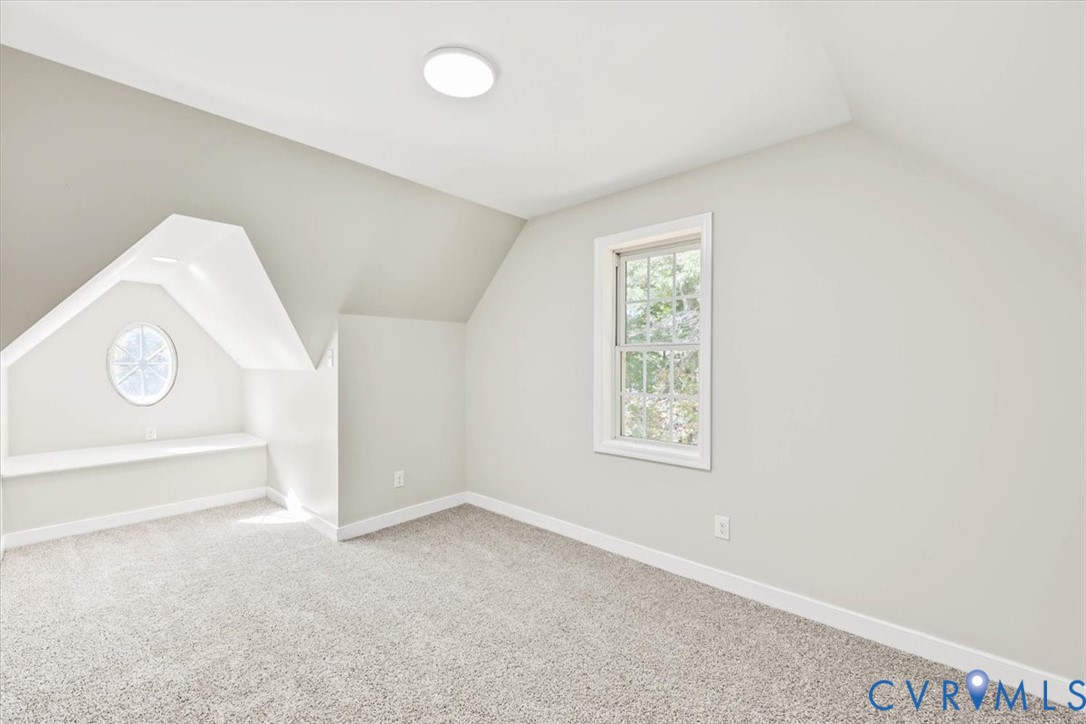 Vaulted ceiling room featuring neutral-toned carpeting, a built-in window seat, an oval accent window, and a double-hung window