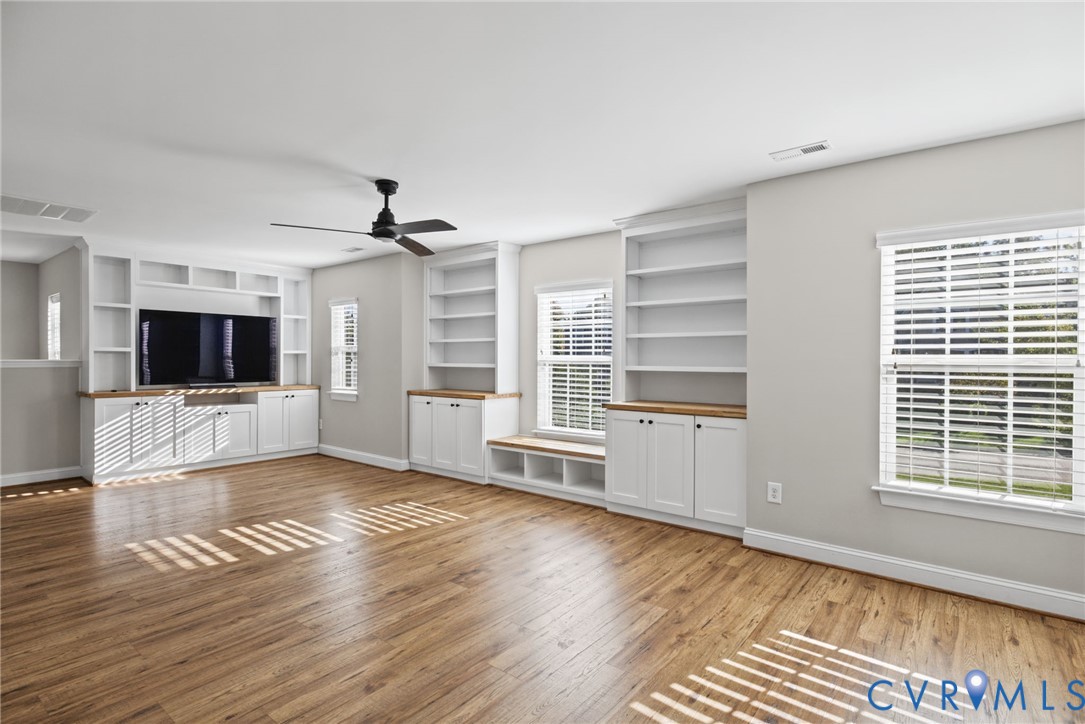 Spacious room featuring wood-finish flooring, custom built-in cabinetry with natural wood-tone surfaces, integrated shelving, a window seat, and a ceiling fan