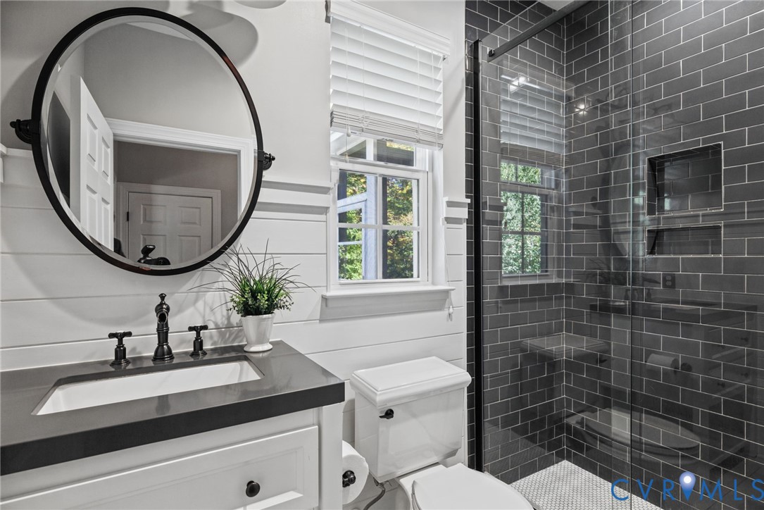 Bathroom featuring white shiplap walls, a vanity with a dark countertop and a circular mirror, a glass-enclosed shower with charcoal subway tile and built-in recessed shelving, and a horizontal-slat window