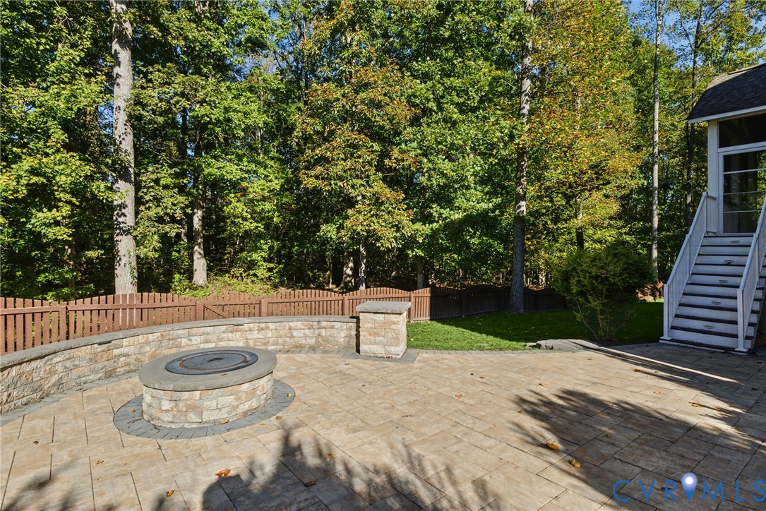 Stone-paver patio featuring a circular masonry fire pit, curved stone-veneer retaining wall, wood-style privacy fencing, and exterior staircase