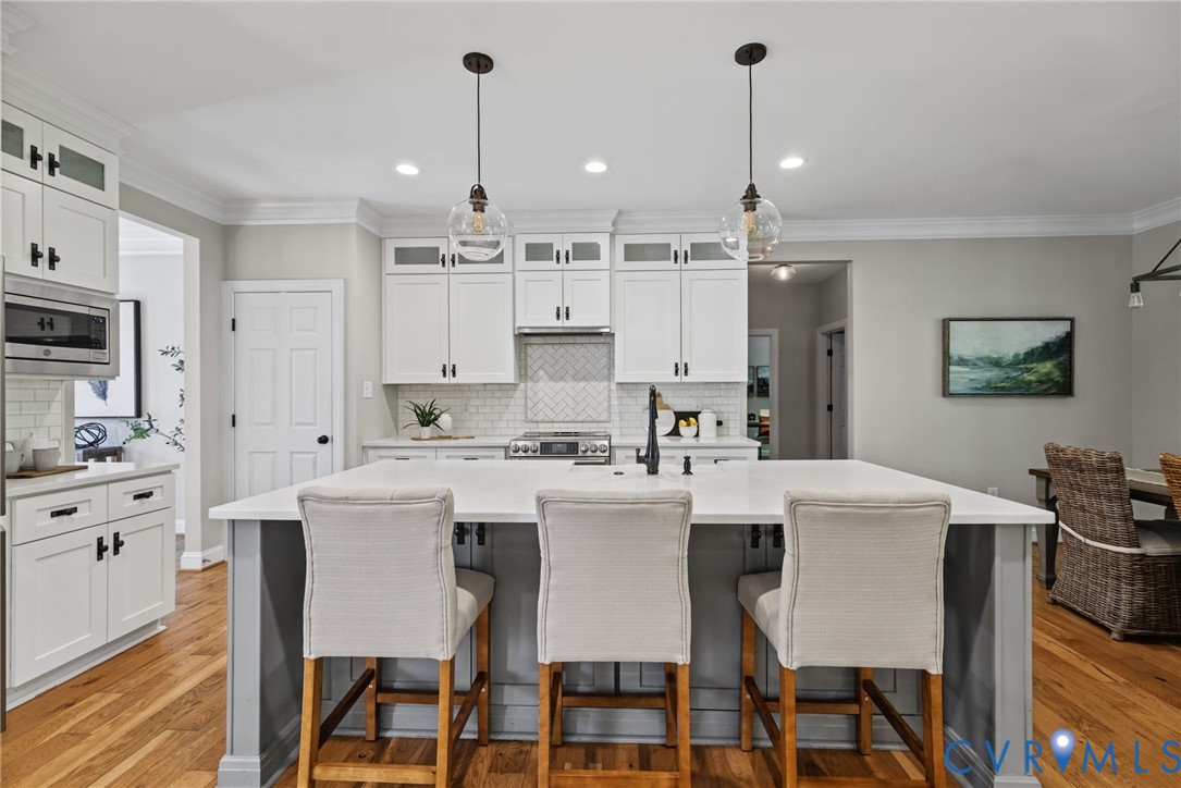 Kitchen featuring a large central island with light-toned solid surface countertops, white shaker-style cabinetry, a herringbone-patterned tile backsplash, and dual glass globe pendant lights over the island