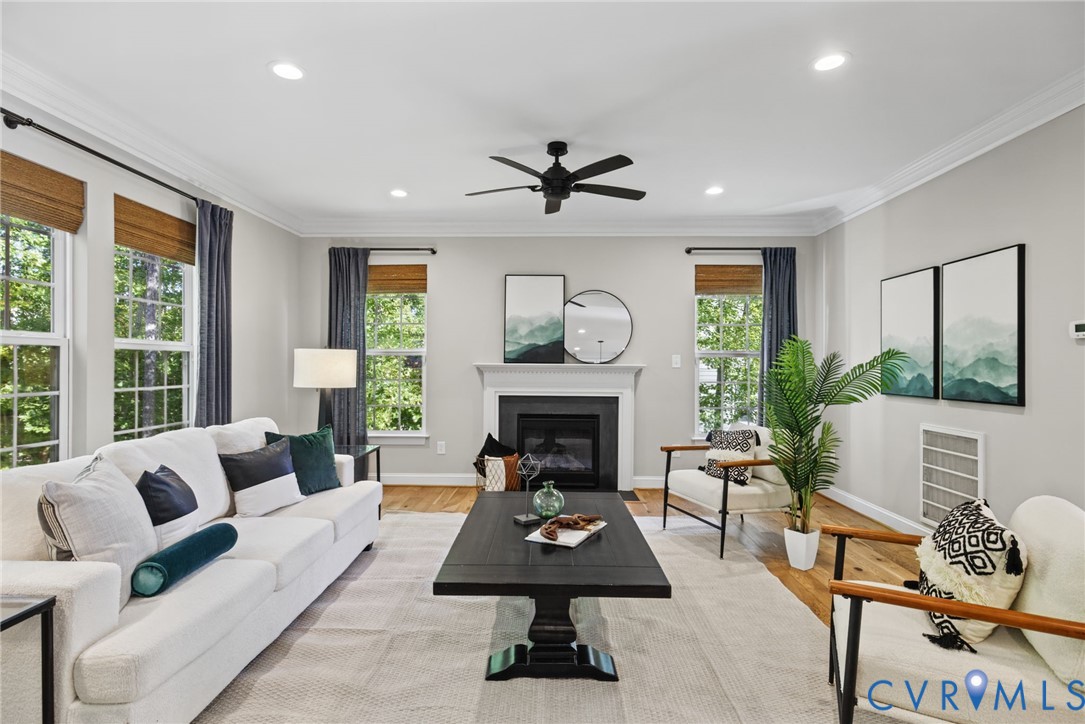 Living area featuring crown molding, wood-finish flooring, a fireplace with a decorative mantel, a central ceiling fan, and multiple windows