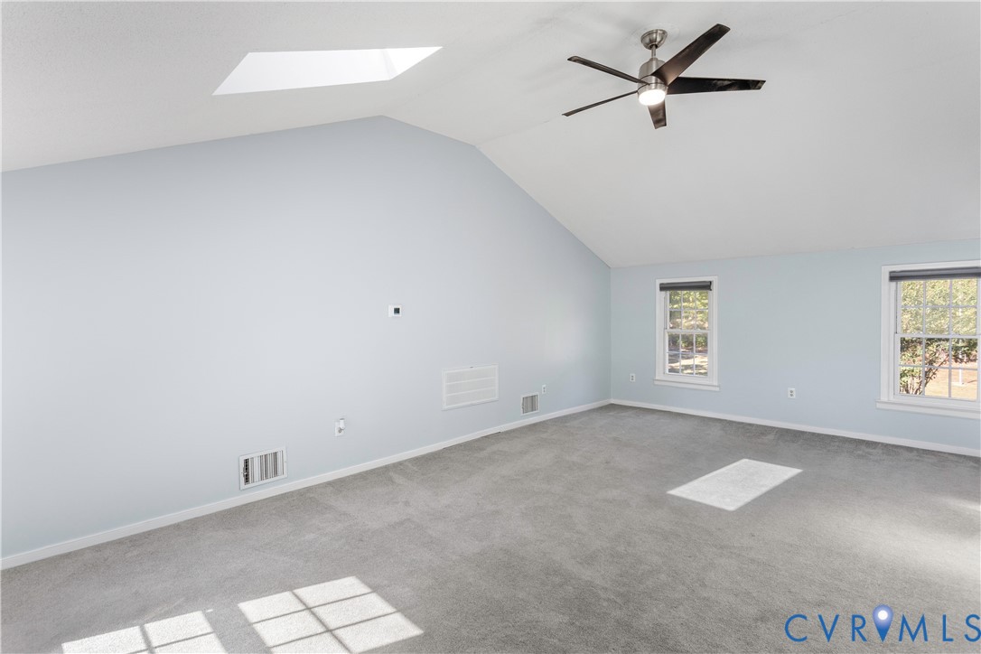 Vaulted ceiling featuring a skylight, carpeted flooring, and a ceiling fan with integrated lighting, complemented by two double-hung windows