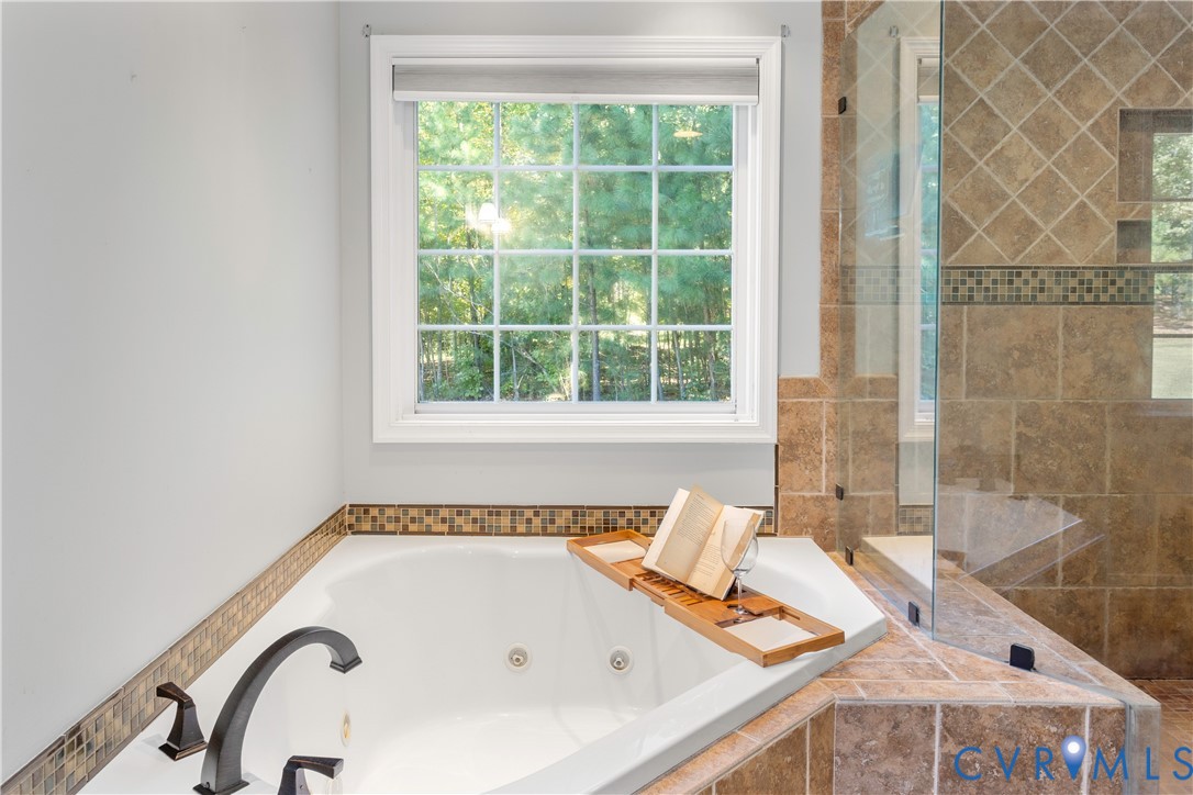 Jetted soaking tub with dark finish faucet, tiled surround, glass-enclosed shower, and decorative mosaic tile accents