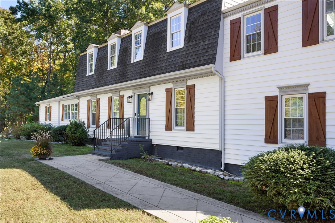 Dutch Colonial exterior featuring white horizontal siding, dark-toned shutters, a gambrel roofline with dormer windows, and a paved walkway leading to metal exterior steps