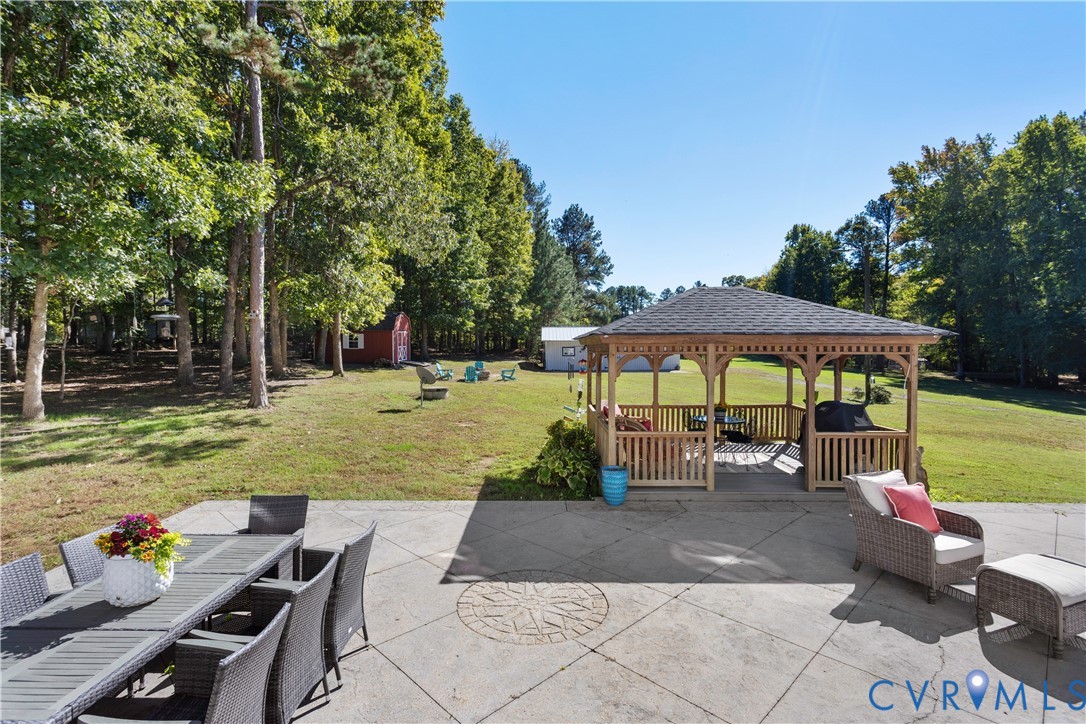 Expansive backyard featuring a concrete patio with an inlaid stone medallion, a wooden gazebo, mature trees, and detached storage structures
