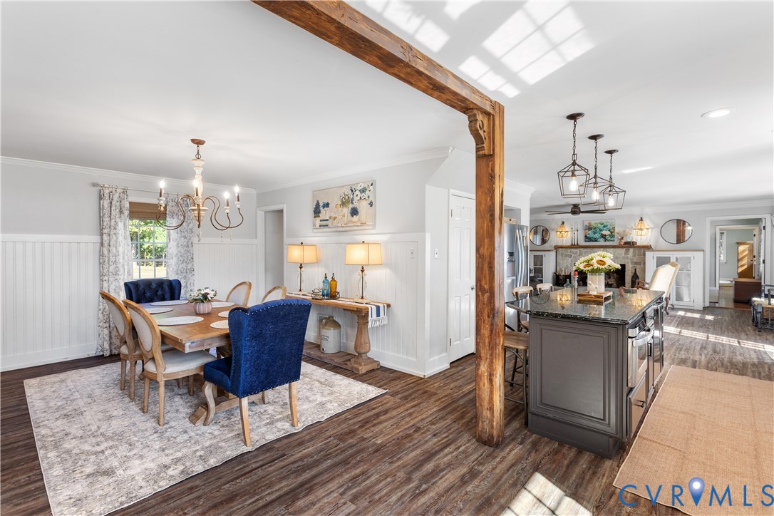 Open-concept interior featuring LTV flooring, a exposed timber structural beam, wainscoting, a kitchen island with dark cabinetry andgranite ountertops, and tiered geometric pendant lighting