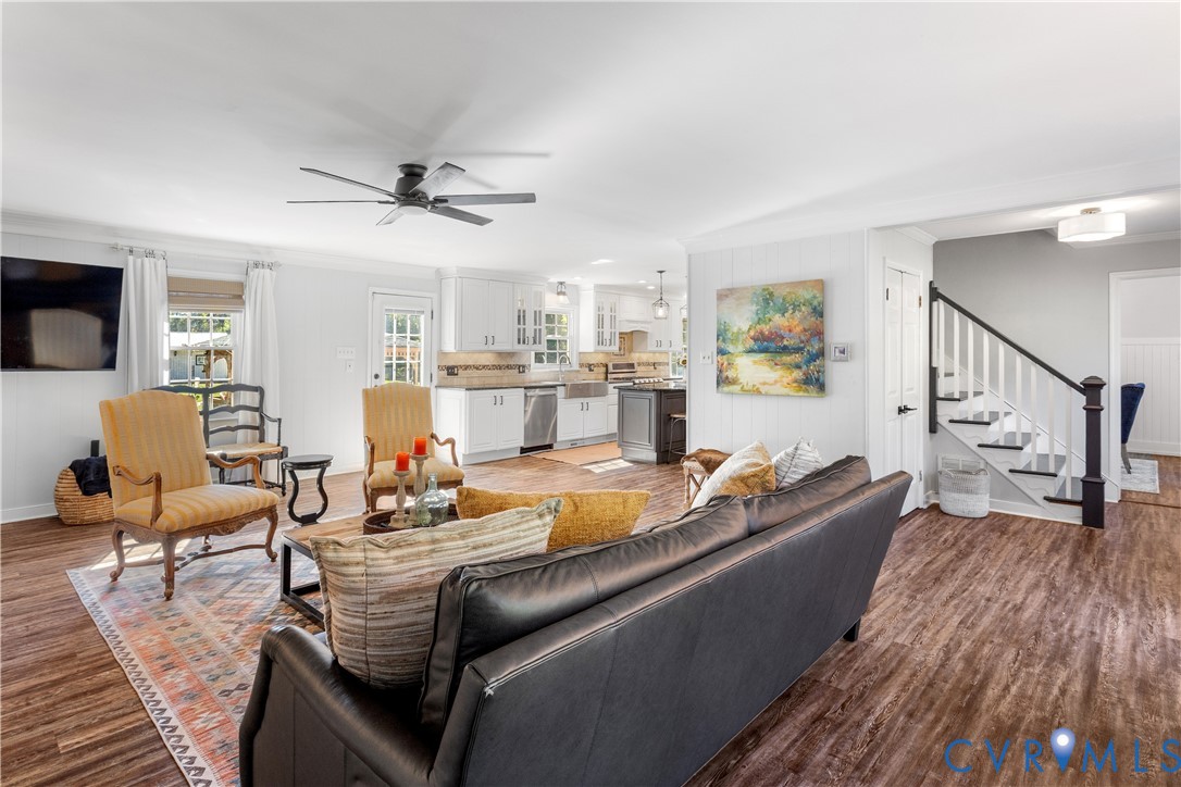Open-concept main living area featuring LTV flooring, a ceiling fan, white cabinetry with stainless steel appliances, a farmhouse-style kitchen sink, and a staircase with a dark wood handrail and white balusters