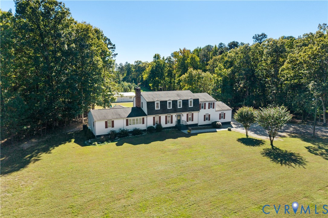 Two-story colonial exterior featuring a brick chimney, gabled dormers, decorative window shutters, and a sprawling lawn surrounded by mature trees