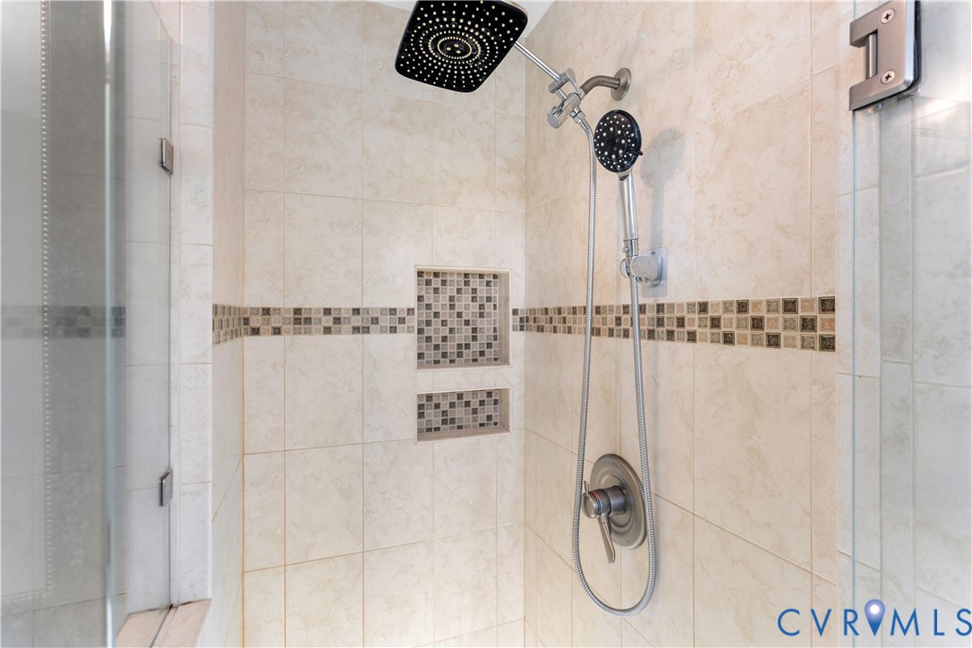 Walk-in shower featuring oversized square rainfall showerhead, adjustable handheld sprayer, neutral-toned wall tiling, decorative mosaic tile inlay, and dual built-in recessed wall niches