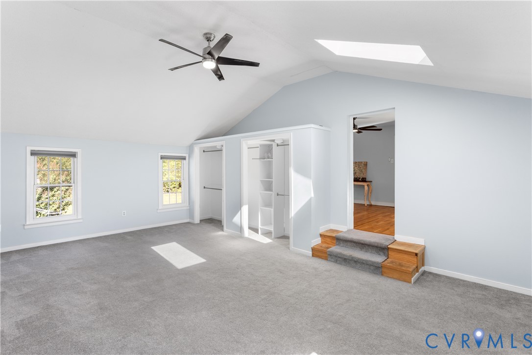 Vaulted ceiling featuring a central skylight, built-in dual-closet system with shelving, integrated ceiling fan, and carpeted flooring with stepped entry